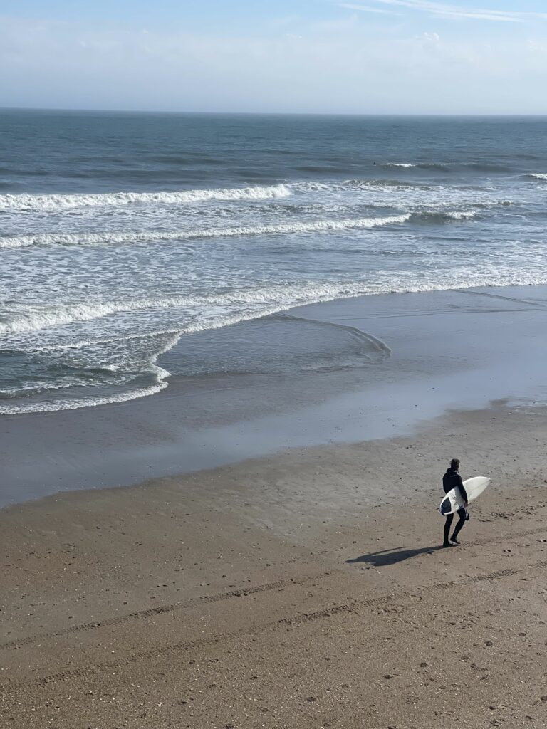 Man walking along Wrightsville Beach with a surfboard. Affordable Outdoor Spots to Explore in Wilmington, NC